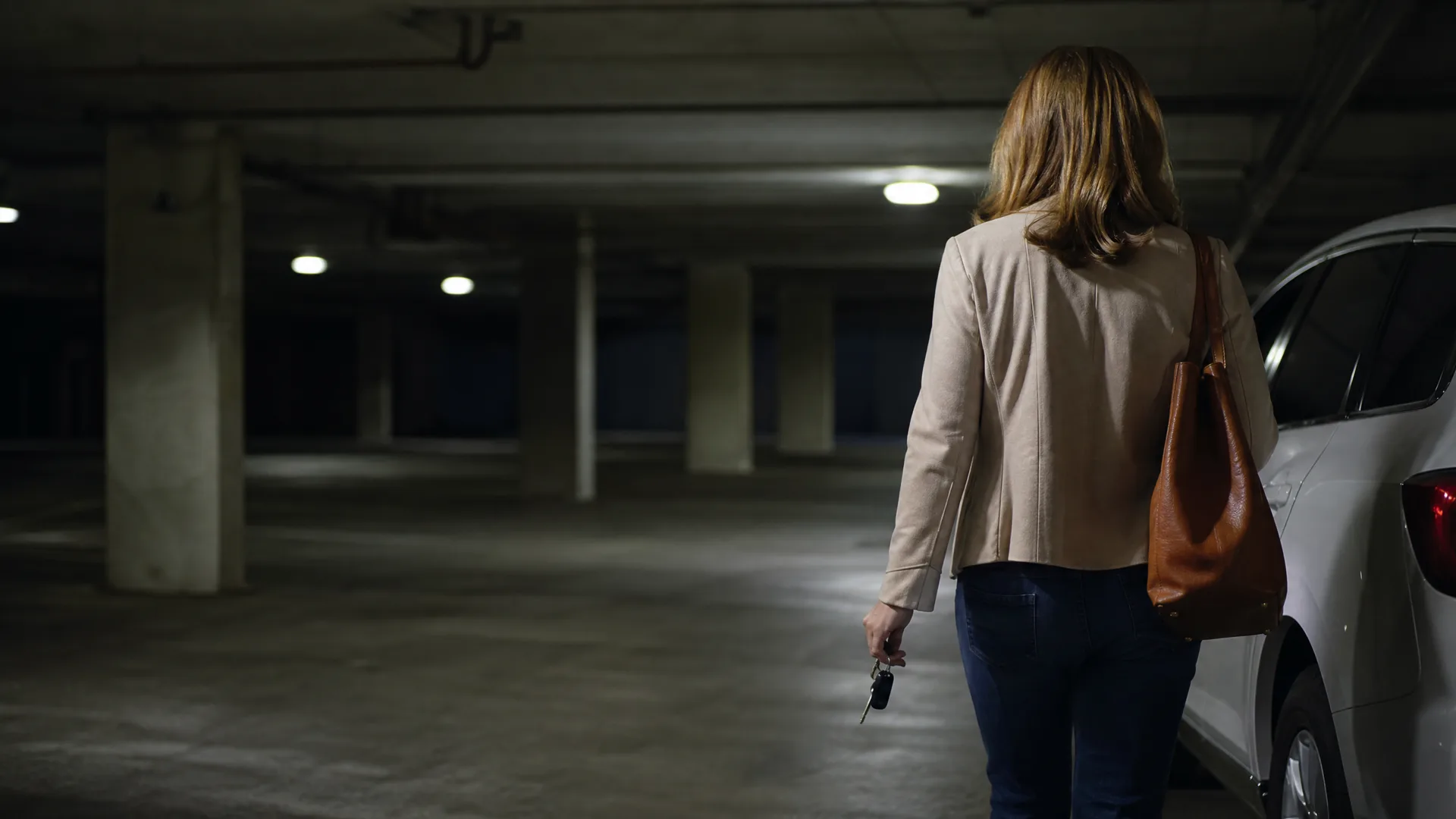 A woman seen from behind in a beige jacket and jeans, carrying a brown shoulder bag and holding car keys, walking in a dimly lit underground parking garage beside a white car.
